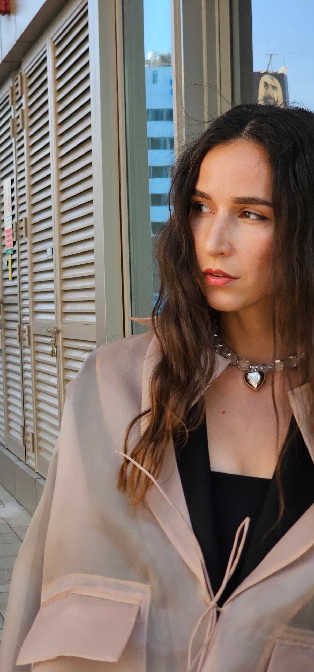Olga Kovaleva in a sheer blush trench coat and a crystal heart choker, looking over her shoulder